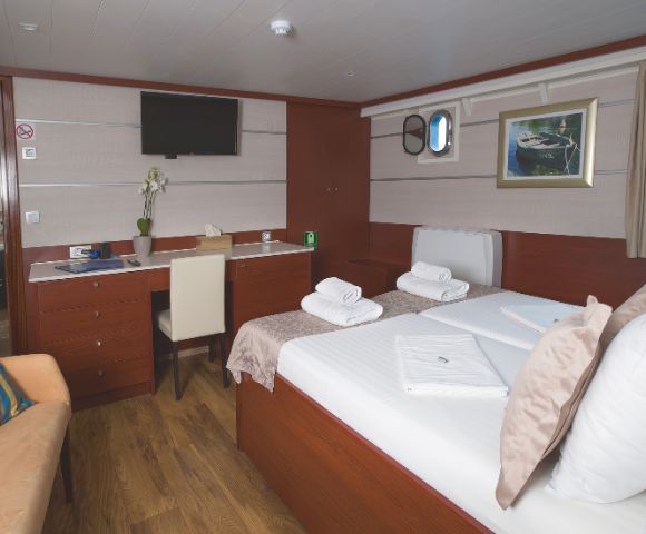 A cozy ship cabin features a neatly made bed with folded towels, a desk with a chair, a wall-mounted TV, and a small window, creating a warm, inviting atmosphere.