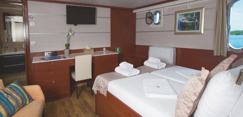 A cozy ship cabin features a neatly made bed with folded towels, a desk with a chair, a wall-mounted TV, and a small window, creating a warm, inviting atmosphere.