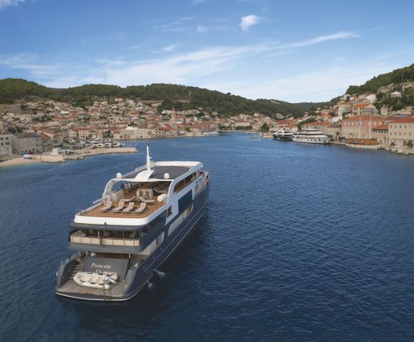 Luxury yacht sailing in a serene bay, surrounded by picturesque coastal town with terracotta roofs and lush hills under a clear blue sky.