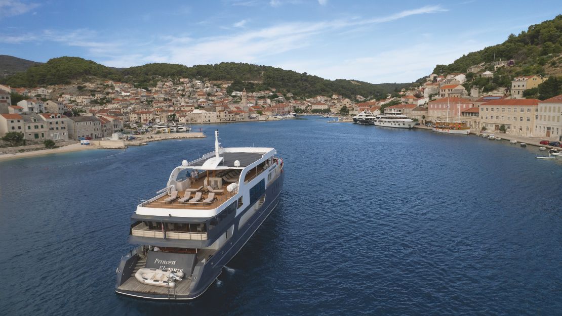 Luxury yacht sailing in a serene bay, surrounded by picturesque coastal town with terracotta roofs and lush hills under a clear blue sky.