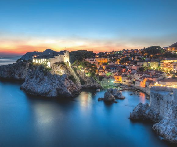 Sunset view of a coastal city with historic stone buildings. A fortress on a cliff overlooks a calm sea, and the skyline glows with city lights. Tranquil and scenic.