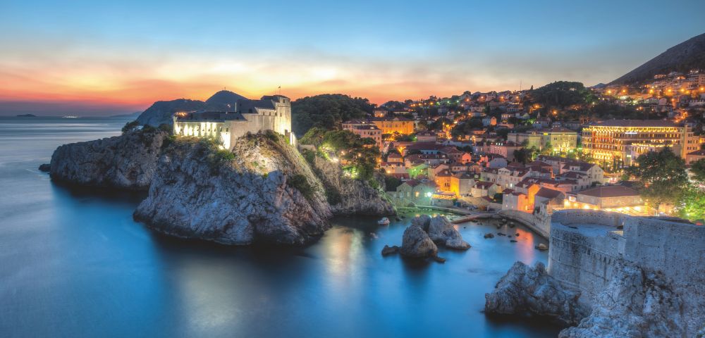 Sunset view of a coastal city with historic stone buildings. A fortress on a cliff overlooks a calm sea, and the skyline glows with city lights. Tranquil and scenic.