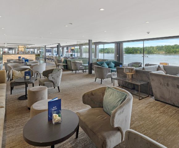 Modern lounge with plush armchairs and sofas, large floor-to-ceiling windows offering a river view. Natural daylight creates a calm, inviting atmosphere.