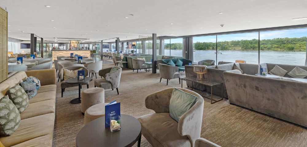 Modern lounge with plush armchairs and sofas, large floor-to-ceiling windows offering a river view. Natural daylight creates a calm, inviting atmosphere.