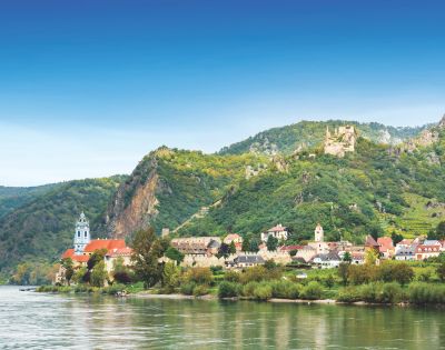 A picturesque riverside village with colorful rooftops sits beneath lush green hills. A blue-domed church tower rises, evoking a serene and quaint atmosphere.