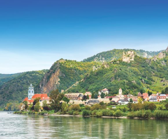 A picturesque riverside village with colorful rooftops sits beneath lush green hills. A blue-domed church tower rises, evoking a serene and quaint atmosphere.