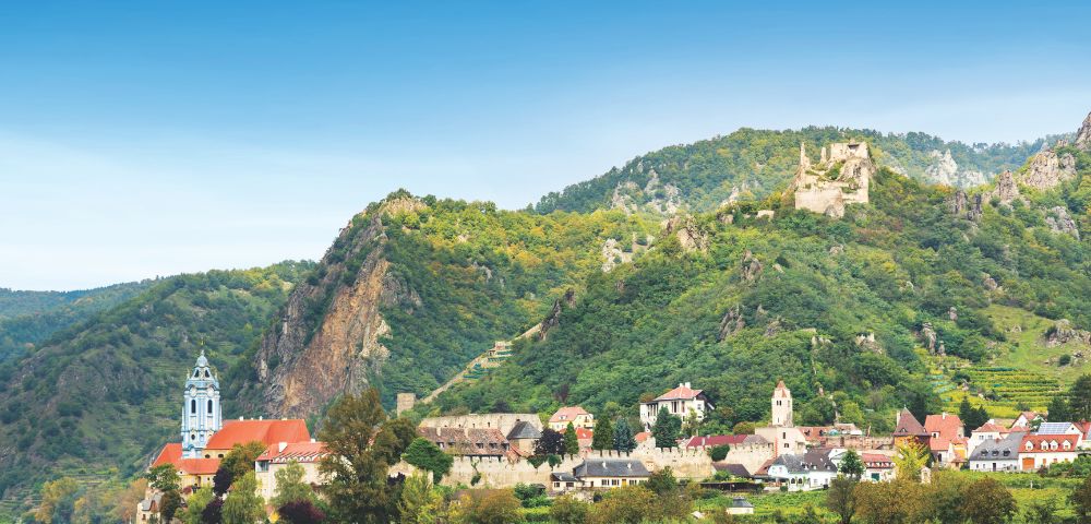 A picturesque riverside village with colorful rooftops sits beneath lush green hills. A blue-domed church tower rises, evoking a serene and quaint atmosphere.