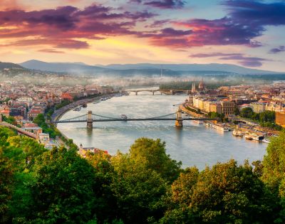 Scenic view of a city with a river winding through, flanked by bridges, under a vibrant sunset sky. Lush greenery in the foreground adds to the beauty.