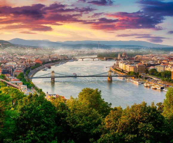 Scenic view of a city with a river winding through, flanked by bridges, under a vibrant sunset sky. Lush greenery in the foreground adds to the beauty.