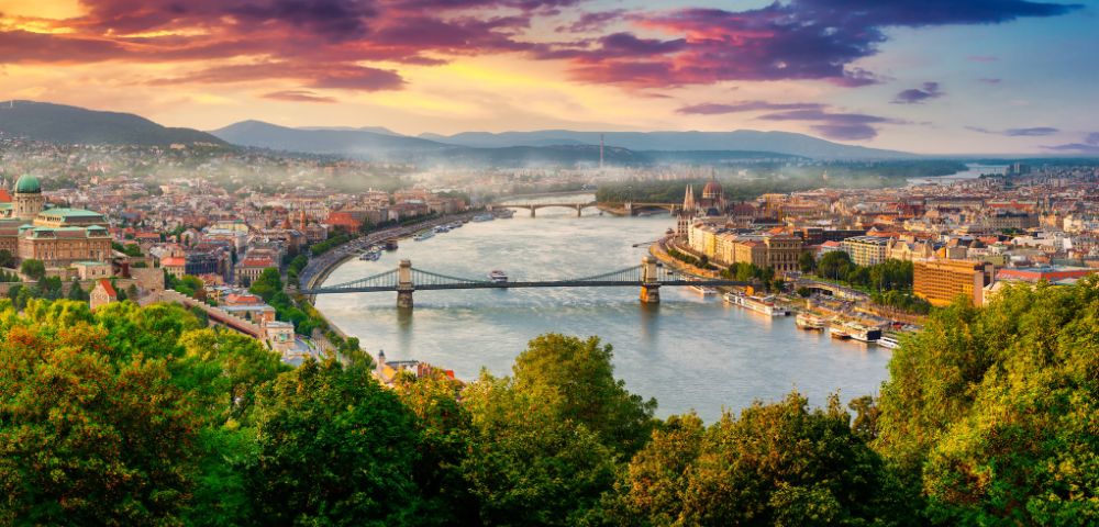 Scenic view of a city with a river winding through, flanked by bridges, under a vibrant sunset sky. Lush greenery in the foreground adds to the beauty.