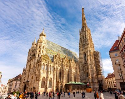 Gothic cathedral with a tall spire and intricate facade under a blue sky. People walk in the square, creating a lively and vibrant scene.