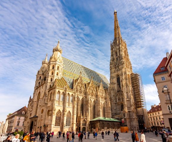 Gothic cathedral with a tall spire and intricate facade under a blue sky. People walk in the square, creating a lively and vibrant scene.