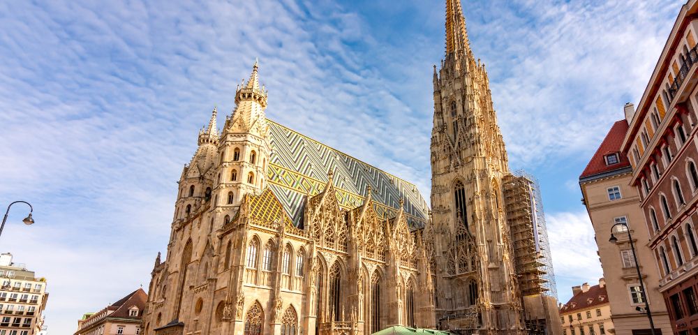 Gothic cathedral with a tall spire and intricate facade under a blue sky. People walk in the square, creating a lively and vibrant scene.