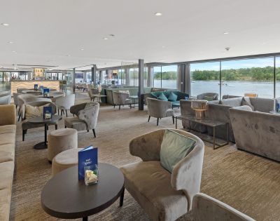 Modern lounge with plush armchairs and sofas, large floor-to-ceiling windows offering a river view. Natural daylight creates a calm, inviting atmosphere.