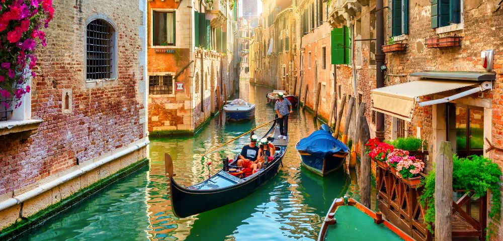 A gondola moves through a narrow canal in Venice, surrounded by historic brick buildings with colorful flowers, green shutters, and boats docked along the water.