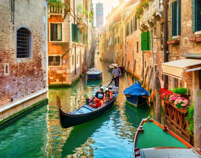 A gondola moves through a narrow canal in Venice, surrounded by historic brick buildings with colorful flowers, green shutters, and boats docked along the water.
