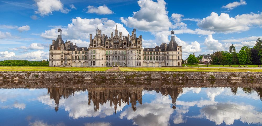 A majestic castle with intricate spires stands against a vibrant blue sky with fluffy clouds. Its reflection shimmers in the calm water below.
