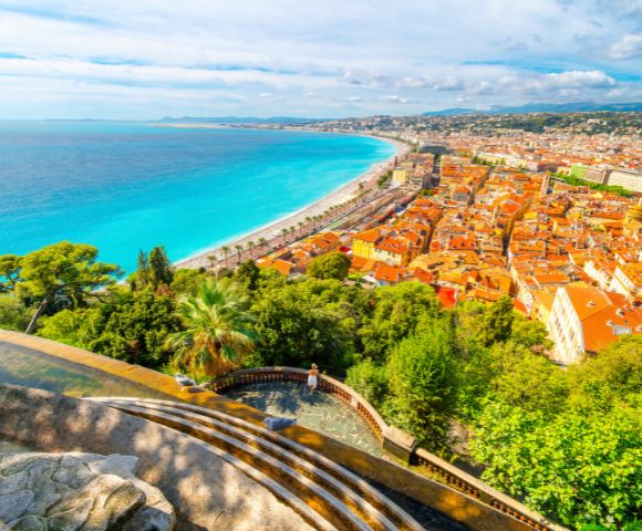 View of Nice, France from above, showing a vibrant blue coastline alongside the city. Orange rooftops, lush greenery, and a bright, airy atmosphere.