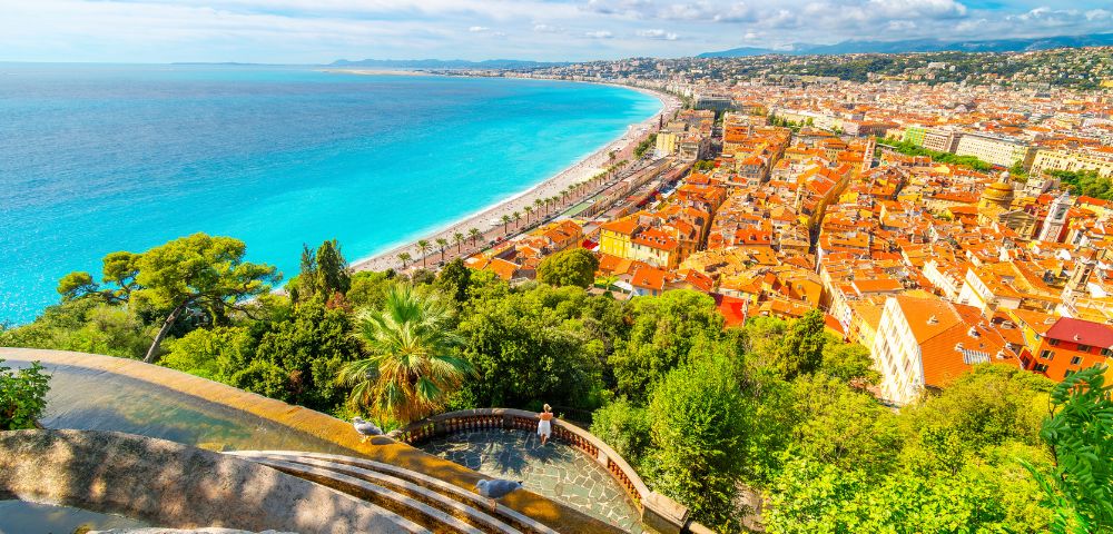 View of Nice, France from above, showing a vibrant blue coastline alongside the city. Orange rooftops, lush greenery, and a bright, airy atmosphere.