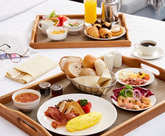 Elegant breakfast spread on wooden trays with croissants, bread, eggs, salmon, deli meats, and coffee on a white tablecloth, conveying a cozy, inviting morning.