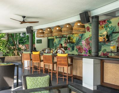 Tropical-themed bar with five wicker pendant lights, floral wallpaper, and a bartender in a white shirt. Green cushions adorn dark rattan chairs.