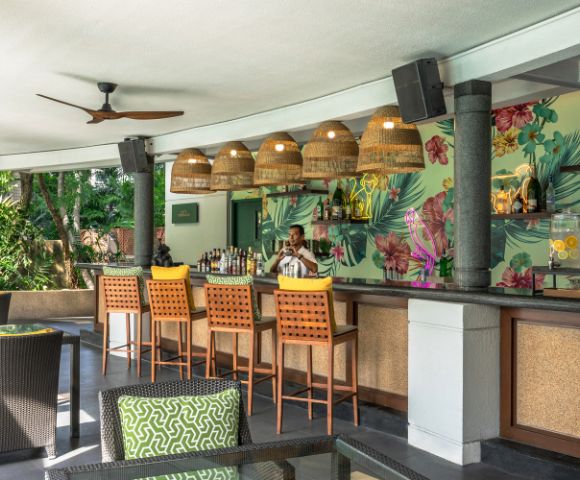 Tropical-themed bar with five wicker pendant lights, floral wallpaper, and a bartender in a white shirt. Green cushions adorn dark rattan chairs.