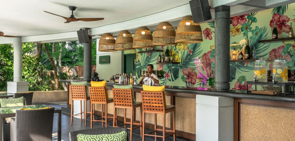 Tropical-themed bar with five wicker pendant lights, floral wallpaper, and a bartender in a white shirt. Green cushions adorn dark rattan chairs.