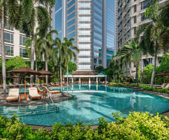 A tranquil hotel pool surrounded by lush palm trees and sun loungers, set against a backdrop of modern high-rise buildings. A relaxing oasis in the city.