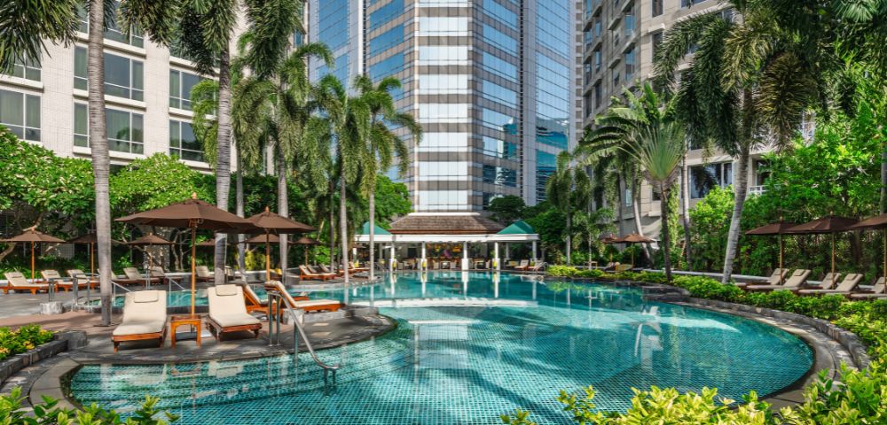 A tranquil hotel pool surrounded by lush palm trees and sun loungers, set against a backdrop of modern high-rise buildings. A relaxing oasis in the city.