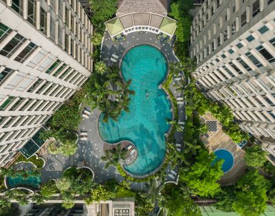 Aerial view of a large, uniquely shaped pool surrounded by two tall buildings, lush greenery, and poolside loungers, conveying a serene, resort-like ambiance.