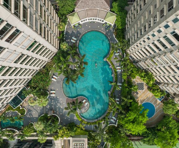 Aerial view of a large, uniquely shaped pool surrounded by two tall buildings, lush greenery, and poolside loungers, conveying a serene, resort-like ambiance.