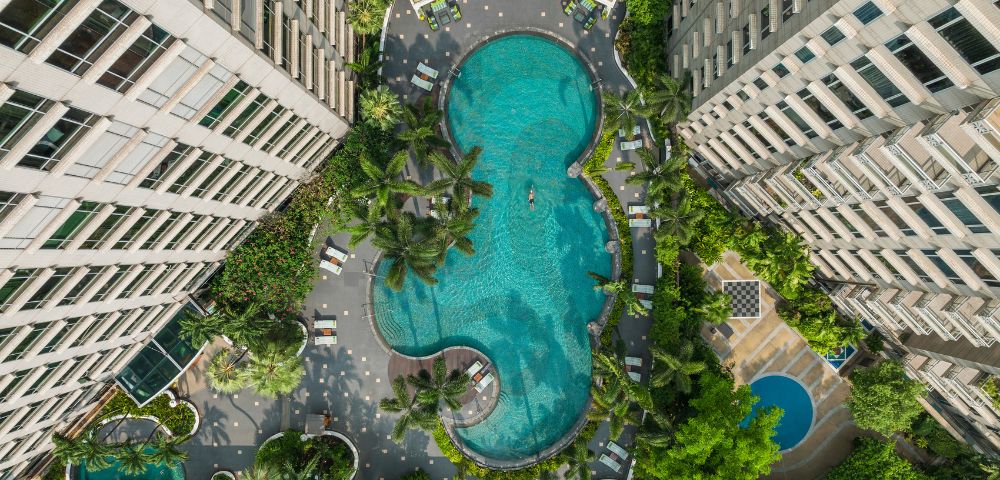 Aerial view of a large, uniquely shaped pool surrounded by two tall buildings, lush greenery, and poolside loungers, conveying a serene, resort-like ambiance.