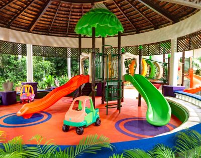 Vibrant indoor playground with red and green slides, colorful plastic car toys, and a circular climbing structure. Bright, cheerful atmosphere.