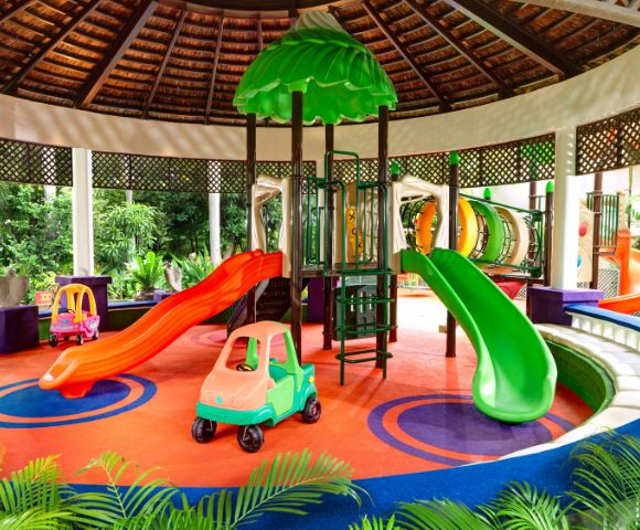 Vibrant indoor playground with red and green slides, colorful plastic car toys, and a circular climbing structure. Bright, cheerful atmosphere.