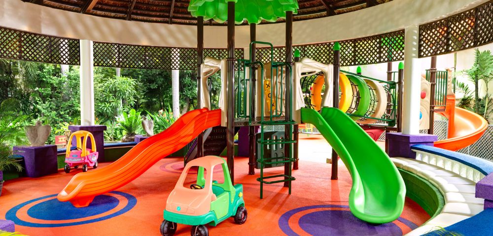 Vibrant indoor playground with red and green slides, colorful plastic car toys, and a circular climbing structure. Bright, cheerful atmosphere.