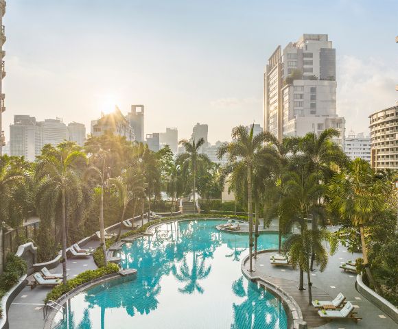Sunlit urban pool surrounded by palm trees, loungers, and high-rise buildings, creating a serene tropical oasis amid a bustling cityscape.