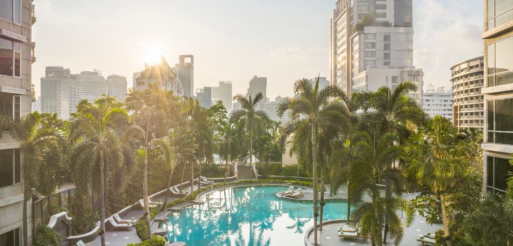 Sunlit urban pool surrounded by palm trees, loungers, and high-rise buildings, creating a serene tropical oasis amid a bustling cityscape.