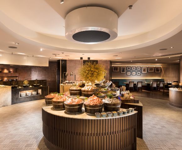 Luxurious buffet restaurant with elegant decor. Central circular table features seafood displays, surrounded by warm lighting and cozy seating.