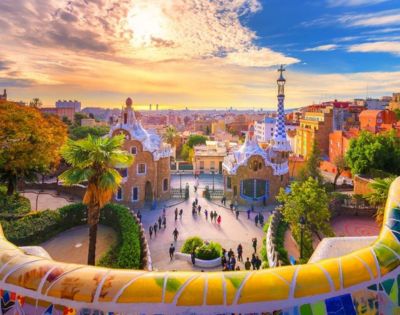 Colorful sunrise over Park Güell in Barcelona, highlighting Gaudí's vibrant mosaic tiles, whimsical architecture, and a lively gathering of visitors.