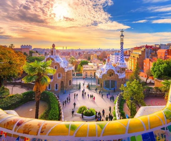 Colorful sunrise over Park Güell in Barcelona, highlighting Gaudí's vibrant mosaic tiles, whimsical architecture, and a lively gathering of visitors.
