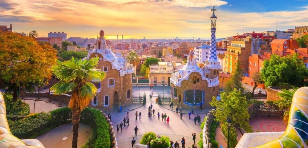 Colorful sunrise over Park Güell in Barcelona, highlighting Gaudí's vibrant mosaic tiles, whimsical architecture, and a lively gathering of visitors.