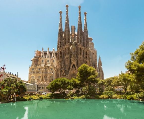The image shows the Sagrada Família basilica in Barcelona, with its intricate spires under a clear blue sky. A small pond and lush greenery are in the foreground.