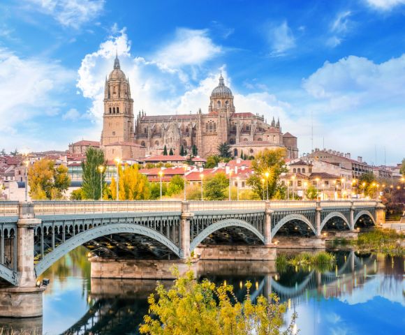 Stunning view of a historic cathedral with twin bell towers overlooking a scenic river and bridge, surrounded by colorful autumn trees under a bright blue sky.