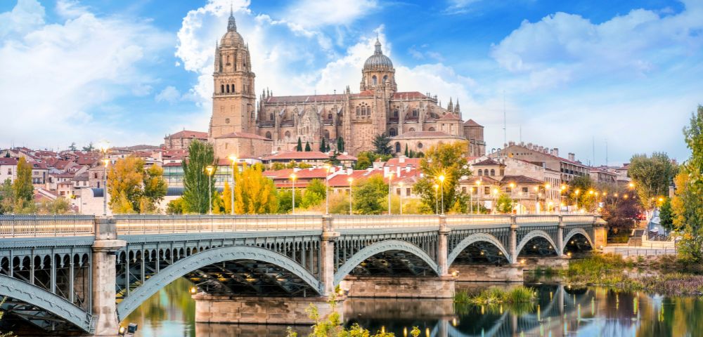 Stunning view of a historic cathedral with twin bell towers overlooking a scenic river and bridge, surrounded by colorful autumn trees under a bright blue sky.