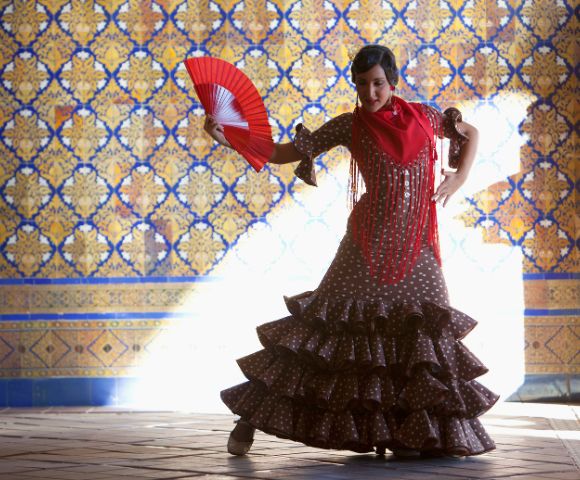 A flamenco dancer in a ruffled polka dot dress, holding a red fan, poses against a patterned tile wall. Sunlight highlights her dramatic stance.