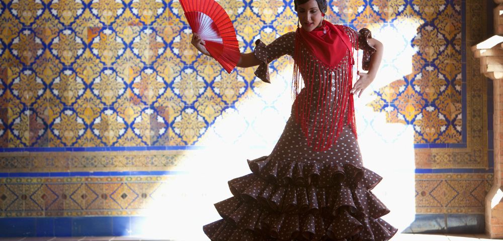 A flamenco dancer in a ruffled polka dot dress, holding a red fan, poses against a patterned tile wall. Sunlight highlights her dramatic stance.