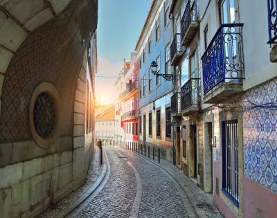 Narrow cobblestone street in Lisbon at sunrise, flanked by colorful buildings with ornate balconies and patterned tiles, creating a serene and picturesque scene.