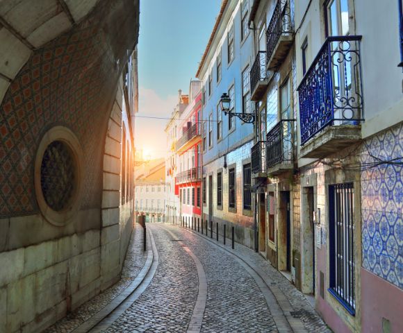 Narrow cobblestone street in Lisbon at sunrise, flanked by colorful buildings with ornate balconies and patterned tiles, creating a serene and picturesque scene.