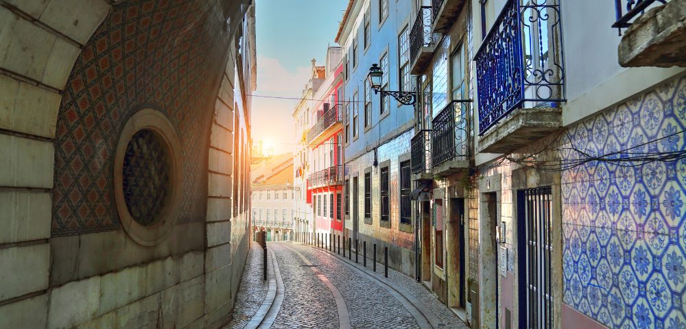 Narrow cobblestone street in Lisbon at sunrise, flanked by colorful buildings with ornate balconies and patterned tiles, creating a serene and picturesque scene.