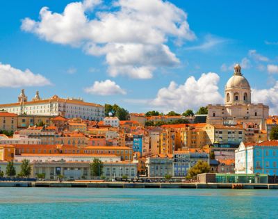 Scenic view of Lisbon with colorful buildings and a domed cathedral under a blue sky with fluffy clouds, reflecting on the calm waterfront.
